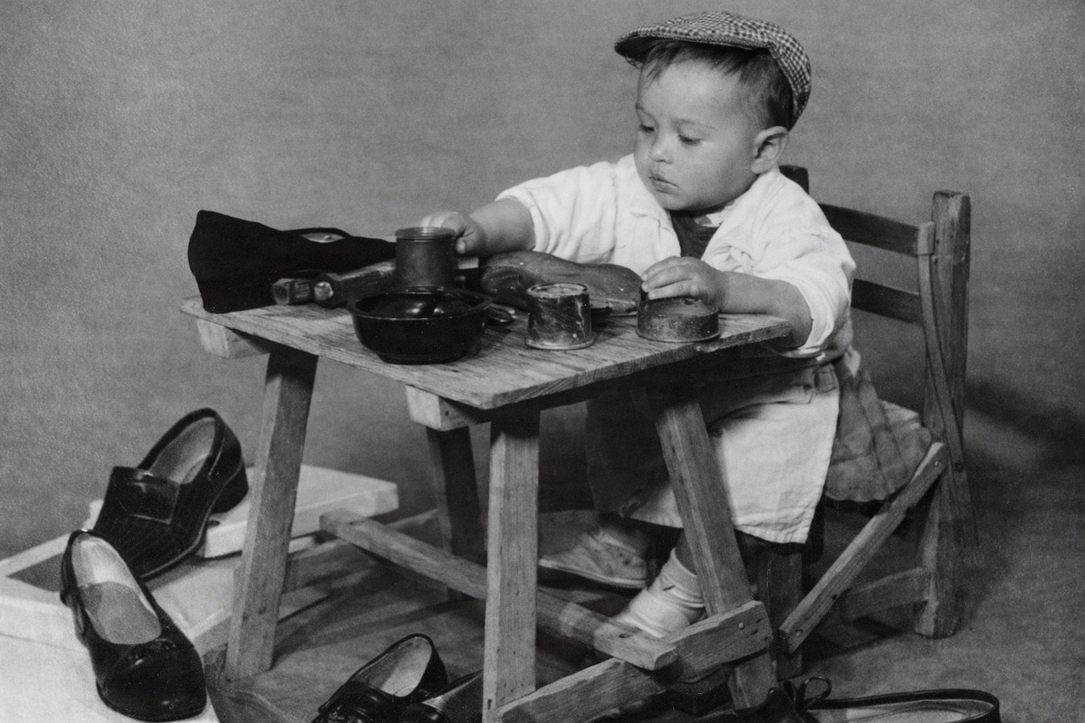 Black and white photo of José Ibáñez Latorre as a child sitting at a small shoemaking bench, surrounded by tools and shoes.